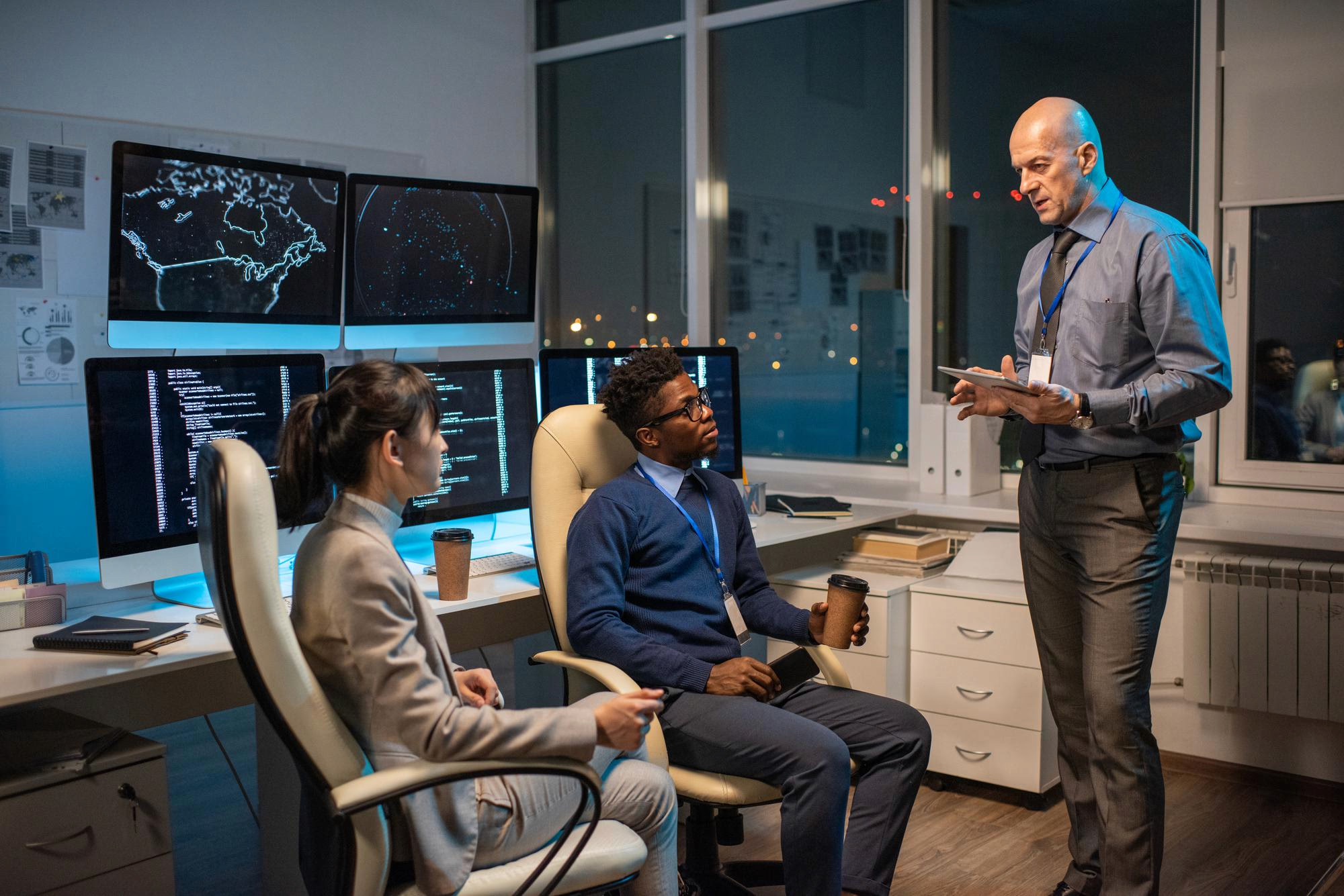 A senior YinkoShield analyst briefing two operators inside the threat-intelligence operations room — multi-monitor world maps and code traces visible in the background.